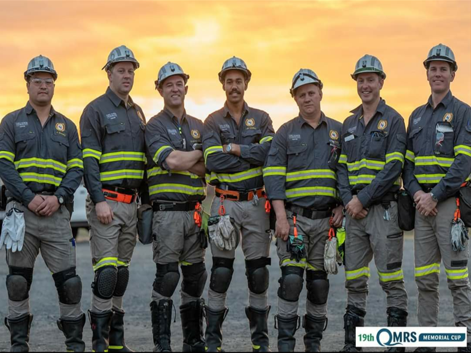 7 men in mining uniforms standing in a row, golden sunset in the background